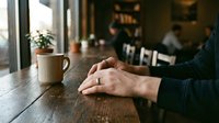 Man's hands with wedding ring resting on rustic wooden table in cozy cafe, coffee mug nearby, warm ambient light, candid lifestyle moment.