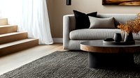 Modern minimalist living room with gray sofa, round wooden coffee table, black and neutral decor elements on textured area rug. Natural light from window creates calm atmosphere.