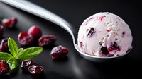 Creamy cranberry ice cream scoop on metal spoon with fresh berries and mint leaves scattered on dark background, frozen dessert close up food photography.