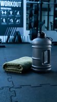 Black water bottle and folded towel on gym floor with workout instruction chart visible in blurred background, representing fitness training and hydration during exercise sessions.