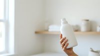 Unisex hand holding white cosmetic bottle in minimalist bathroom setting with natural light and wooden shelves for skincare product marketing.