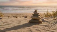 Zen stone stack on sandy beach at golden hour sunset with ocean waves and dune grass creating peaceful meditation atmosphere for wellness and mindfulness concepts.