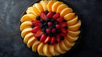 Fresh fruit tart with arranged peach slices, strawberries, blackberries and raspberries on dark chocolate ganache in round baking dish on black background.
