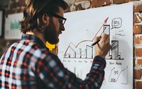 Caucasian bearded businessman in plaid shirt drawing financial growth chart on whiteboard. Business analytics and strategy planning concept shown through hand-drawn graphs.