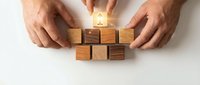 Male hands arranging wooden blocks with glowing house icon on top, representing property investment, real estate planning, home building, and financial growth strategy.