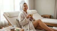 Happy young woman in bathrobe and towel enjoying morning tea in bright bedroom after spa treatment, relaxing at home with healthy lifestyle routine.