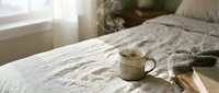 Steaming ceramic mug of hot coffee on a cozy linen bed with a book and knit blanket in soft morning light.