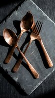 Copper flatware set with hammered handles arranged on slate tile against dark wood surface, casting dramatic shadows for dining and tableware styling.