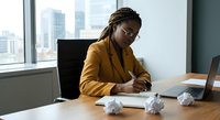Professional African American businesswoman in yellow blazer writing notes at modern office desk with laptop and crumpled papers, city skyline visible through window.