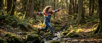 Young blonde girl jumping over forest stream with arms spread wide, exploring nature adventure in mossy woodland setting for outdoor childhood concepts.