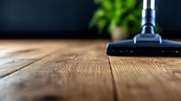 Modern vacuum cleaner head on wooden floor with blurred green plant in background, close-up view showing texture and grain of hardwood flooring.