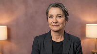 Professional mature businesswoman with gray hair in dark suit smiling confidently in modern office setting with warm lighting for corporate presentations.