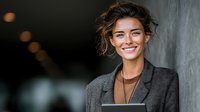 Professional businesswoman with curly hair smiling confidently while holding tablet in modern office environment for corporate presentations and meetings.
