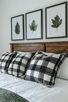 Cozy bedroom interior with wooden headboard, plaid pillows and botanical leaf artwork in black frames creating modern farmhouse decor style.