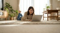 Woman of East Asian descent lying on carpet using laptop at home, relaxed casual work environment.