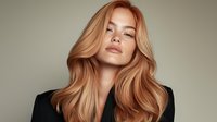 Young woman with flowing strawberry blonde hair wearing black blazer against neutral background, professional beauty portrait showcasing natural makeup and hair styling.