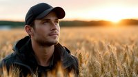 Young Caucasian farmer in baseball cap standing in golden wheat field at sunset, agricultural worker contemplating harvest season in rural countryside.
