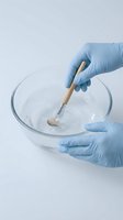 Laboratory technician hands in blue gloves mixing white powder substance with wooden spatula in glass bowl for scientific research and testing.