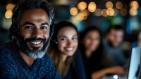 Middle-aged South Asian male with dark beard and salt-and-pepper hair wearing headphones, smiling at camera in modern office setting with diverse colleagues and bokeh lighting in background.