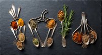 Colorful spices and herbs arranged in vintage wooden spoons on dark slate background. Culinary seasoning collection for cooking and food preparation.
