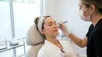 Professional cosmetologist applying eyebrow treatment to relaxed female client in modern beauty salon with natural lighting and clinical atmosphere.
