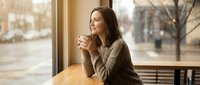 Young brunette woman enjoying hot beverage at cafe window table during winter day, perfect for lifestyle and hospitality marketing materials.