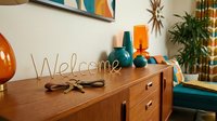 Retro mid-century modern living room with wooden sideboard, gold wire welcome sign, keys, and colorful teal and orange decor accents.