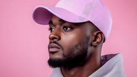 Young African American man wearing pink baseball cap and gray hoodie against pink background, profile portrait showing confidence and style.