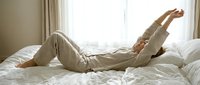Young Caucasian woman in cozy linen pajamas stretching on white bed after waking up, enjoying relaxed morning routine in bright bedroom.