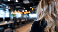 Blonde woman with wavy hair in modern office space with blurred background and bokeh lights, back view. Professional business environment with soft focus.