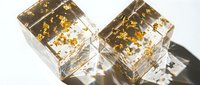 Luxury gold leaf ice cubes with floating flakes in clear liquid, casting shadows on white surface for premium beverage and cosmetic product photography.