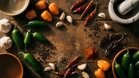 Assorted spices and fresh ingredients arranged on rustic wooden table including chili peppers, garlic, turmeric powder and cooking utensils for culinary preparation.