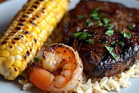 Grilled surf and turf dinner featuring juicy steak, seasoned shrimp, charred corn on cob with butter, served over fluffy white rice and garnished with fresh herbs.