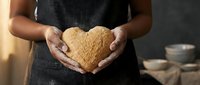 Woman baker holding heart-shaped dough in cupped hands wearing dark apron in rustic kitchen setting with warm lighting and pottery in background.
