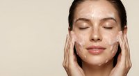 Young woman applying facial cleanser with closed eyes and gentle smile, hands massaging white cream into skin for daily skincare routine and beauty care.