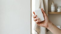 Woman's hand holding blank white product box against neutral bathroom wall with wooden shelf and toiletries for packaging mockup and branding.