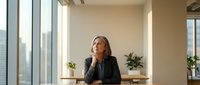 Professional businesswoman in contemplative pose at modern office desk with city view through large windows during golden hour lighting.