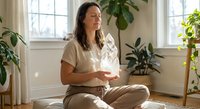 Young woman meditating peacefully in bright living room surrounded by green plants, practicing mindfulness and wellness at home for mental health and relaxation.