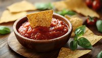 Fresh tomato salsa dip in brown ceramic bowl with crispy tortilla chips, garnished with basil leaves on wooden board, selective focus on appetizer.
