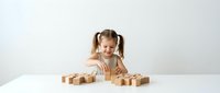 Happy young girl with pigtails playing with wooden building blocks on white table, educational toy development activity for preschool learning and creativity.