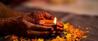 Hands with intricate henna patterns holding glowing oil lamp surrounded by orange marigold petals during traditional celebration ceremony.