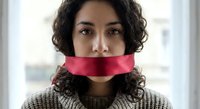 Young Hispanic woman with curly hair and red tape over mouth symbolizing censorship, silence, and freedom of speech restrictions in modern society.