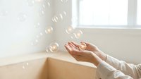 Woman with fair skin wearing a light long-sleeved shirt catching floating soap bubbles near a bright window, symbolizing joy, lightness, and carefree moments for wellness and lifestyle content.