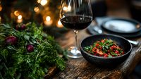 Elegant table setting with red wine glass, fresh herbs and gourmet meal served in black bowl on rustic wooden surface. Moody restaurant atmosphere with bokeh lights.