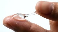 Close-up of a person's fingers holding a clear liquid droplet on a needle or syringe tip against a white background, suitable for medical, scientific, pharmaceutical, and healthcare applications.