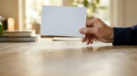 Senior man with medium skin tone holding blank white card on wooden table in home office setting, suitable for business messaging and promotional campaigns.