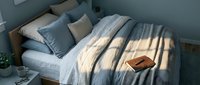 Cozy bedroom interior with unmade bed, soft morning sunlight streaming through window blinds creating shadow patterns on white bedding and pillows.