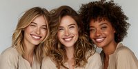 Diverse group of three smiling women with different hair textures wearing neutral beige tops against light background, representing friendship and inclusivity.