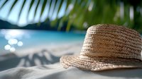 Straw sun hat on sandy tropical beach with turquoise ocean water and palm leaves, summer vacation concept with natural sunlight bokeh effect.