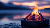 Glowing campfire in portable fire pit against blurred sunset lake background creates cozy outdoor ambiance with burning logs and orange flames.